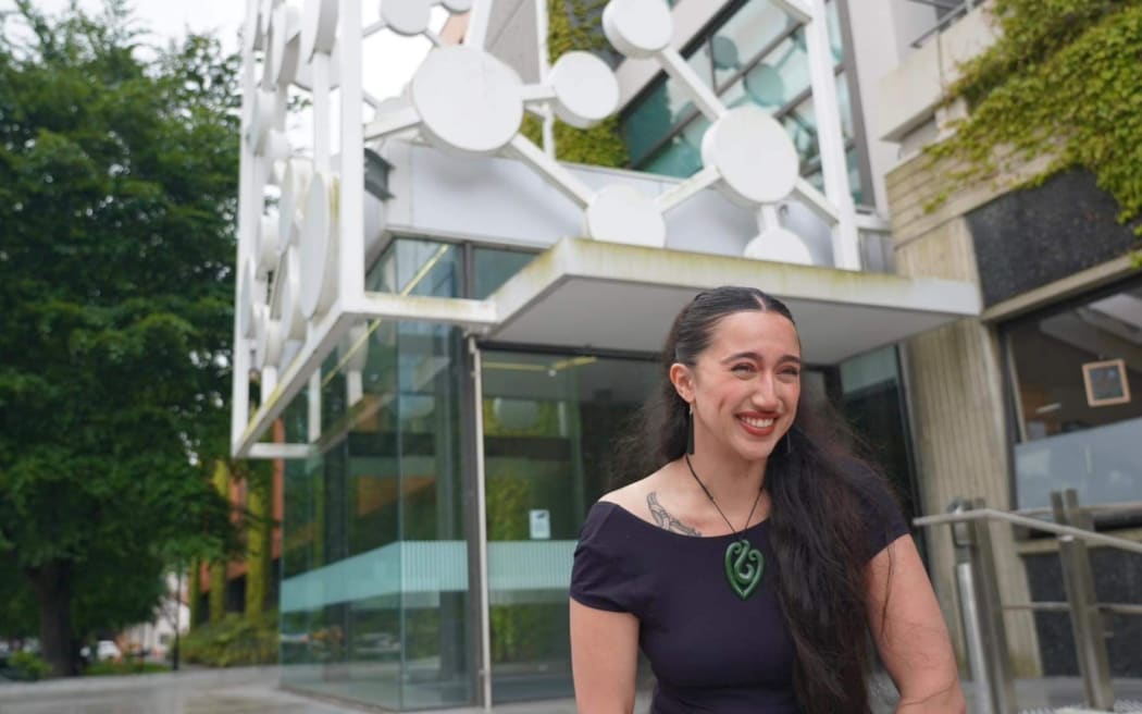 A woman with long hair and a big smile wearing a pounamu pendant in front of a science building with a white molecule design on its facade.