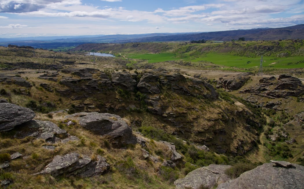 A landscape photo taken from high on a hill showing dry brown hills studded with rocky tors, patches of scrub, and a green grass valley floor and lake in the distance.