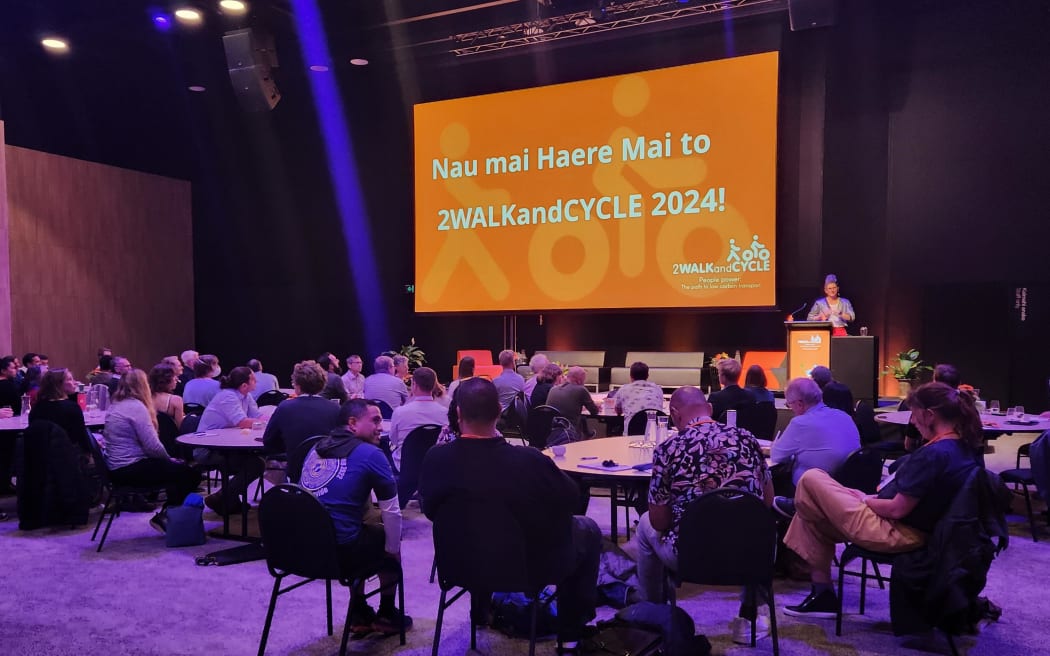 People sitting at round tables at a cycling and walking conference. There is purple lighting and a large screen showing a welcome message with an orange background.