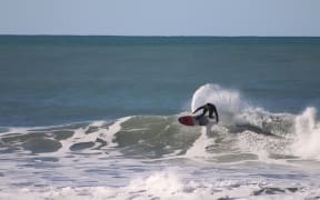 Deejay surfs a wave and throws out spray behind him. It is a blue-sky, sunny day.