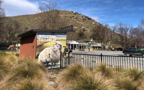 A picture of the Tarras township. In the background are small buildings and behind them, hills. The side of a public bathroom is painted with a large sheep and has text reading "WELCOME TO TARRAS".