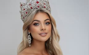 Georgia smiles at the camera. She is wearing a white off-the-shoulder gown and a large silver and red crown.