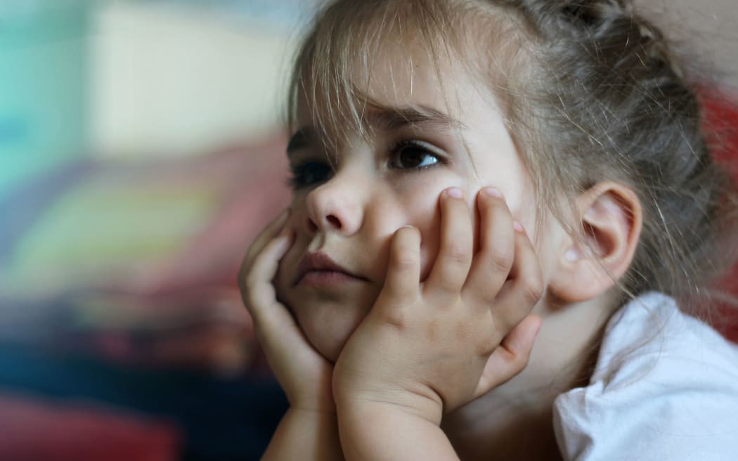 A photo of a little girl lying on the sofa with hands on chin watching the television and looking worried