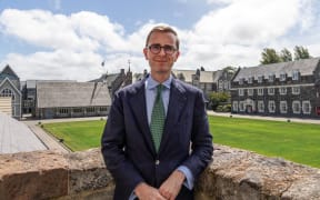 Jeremy stands against a raised stone wall with the Christ's College grounds behind him. He is wearing a suit with a green tie. He smiles.