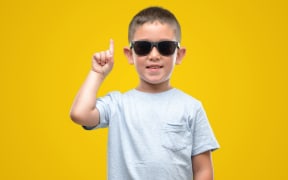 Dark haired little child wearing sunglasses surprised with an idea or question pointing finger with happy face, number one