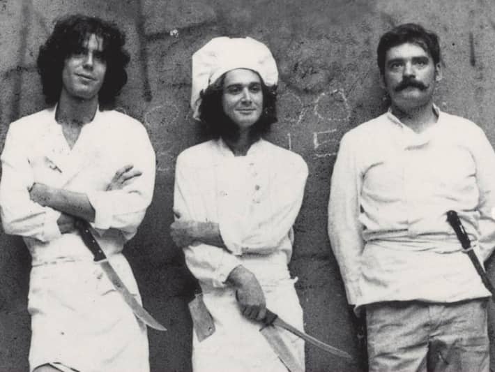 Three men wearing white aprons and carrying knifes are lined up against a wall in a black and white photo.