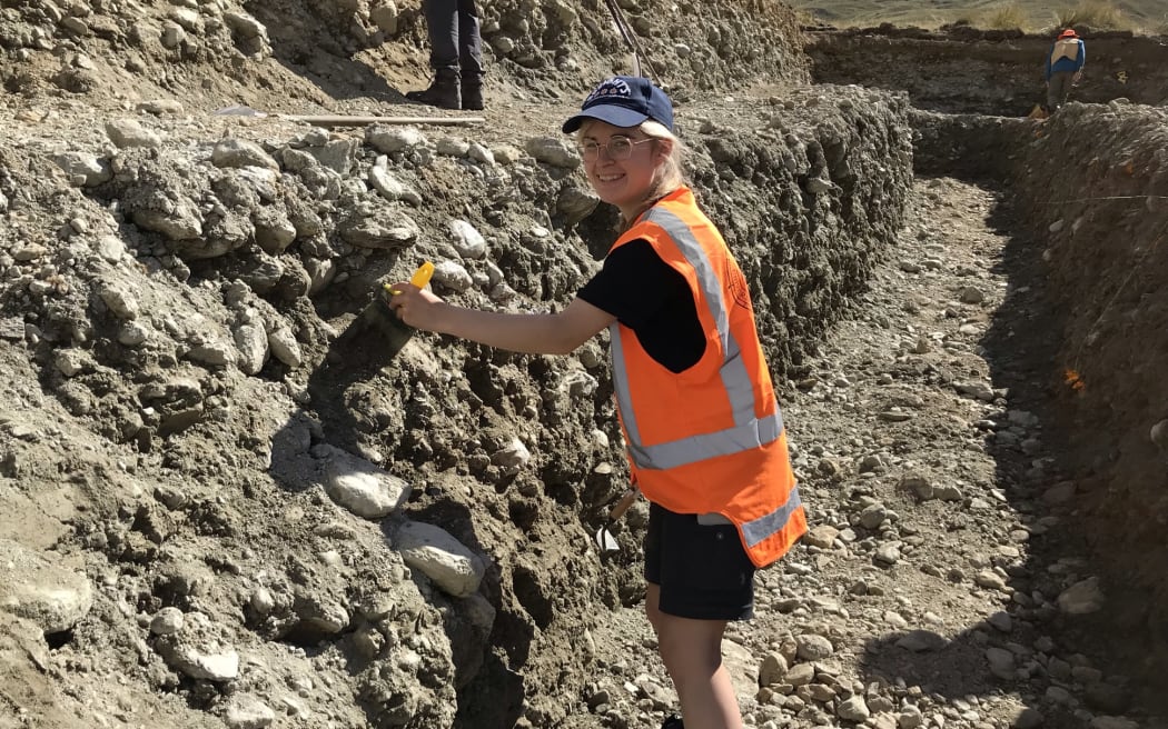 Geology Masters student Ashleigh Vause cleans off the bottom of the trench. Ashleigh is wearing an orange high vis vest and a blue speights baseball cap. She has a paint brush in her hand as she brushes soil off the side of the trench, she is turned towards the camera and smiling.