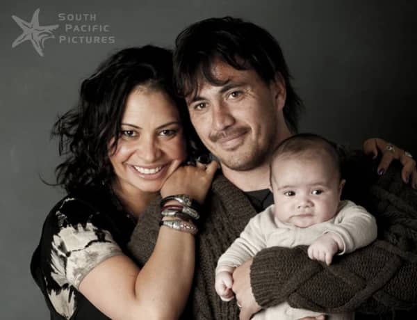 Two young parents smile for the camera with the dad holding a young baby.