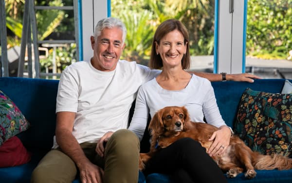 Chris Eggleston (left) and Ali Swarbrick  (right) sitting on their living room couch with their dog Baz. Painter tape edges the windows behind them indicating the renovation their home is currently having.