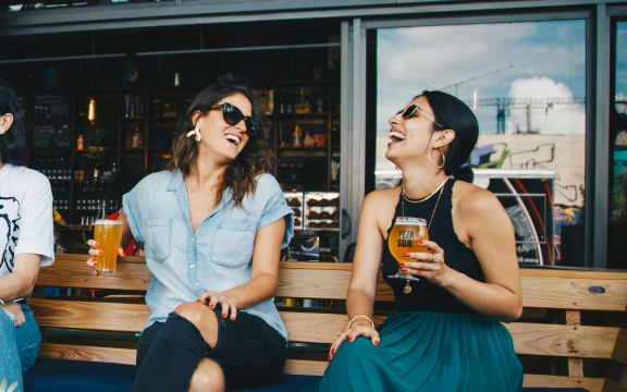 Two women in sunglasses sit outside at a bar laughing and drinking beer.