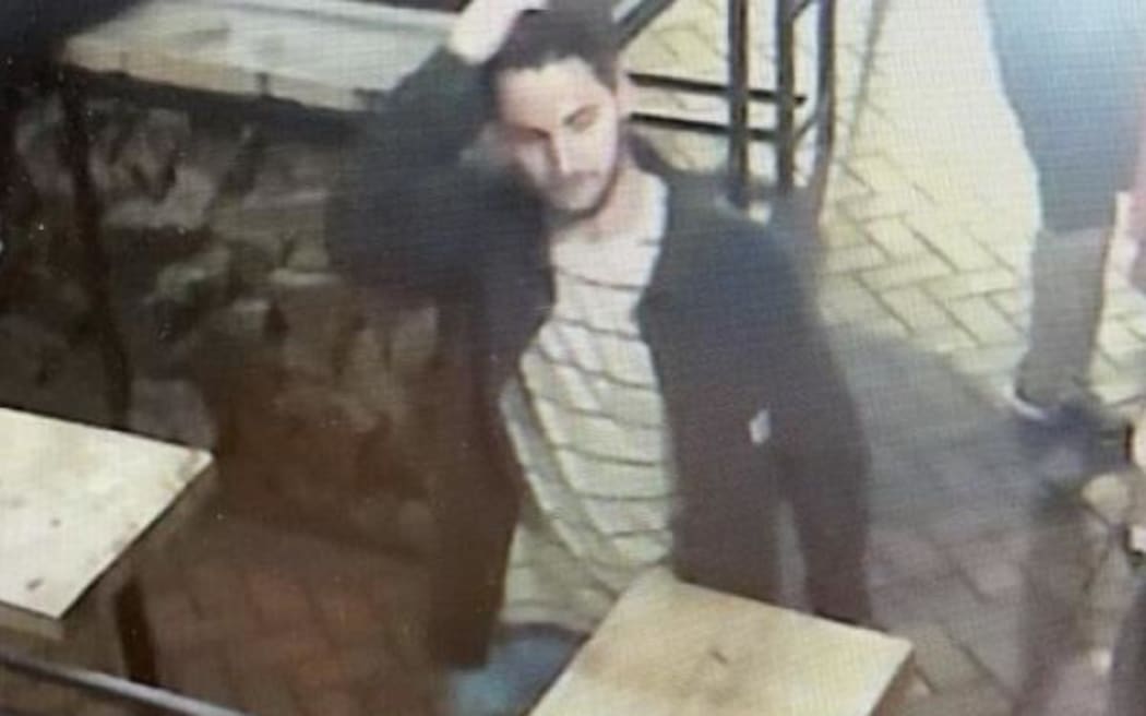 A man wearing a striped white top with a dark coloured jacket walks between tables. He is running his right hand through his-- dark coloured hair.