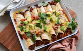 A rectangular dish of tofu and black bean enchiladas topped with avocado and coriander leaves.