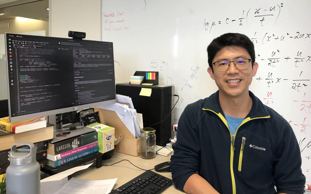 A man sits in front of a computer and a whiteboard, smiling at the camera. The computer screen is black and filled with code, the whiteboard is covered with mathematical equations.