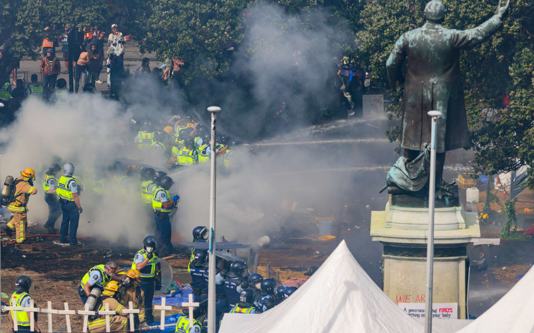 Police stand in the hot embers of the first fire to aim hoses at the second.