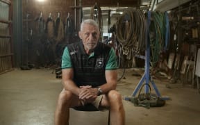 Mark Graham sits on a chair in a warehouse, looking intently at his interviewer to the right of the camera. He wears a green tshirt and a black vest.