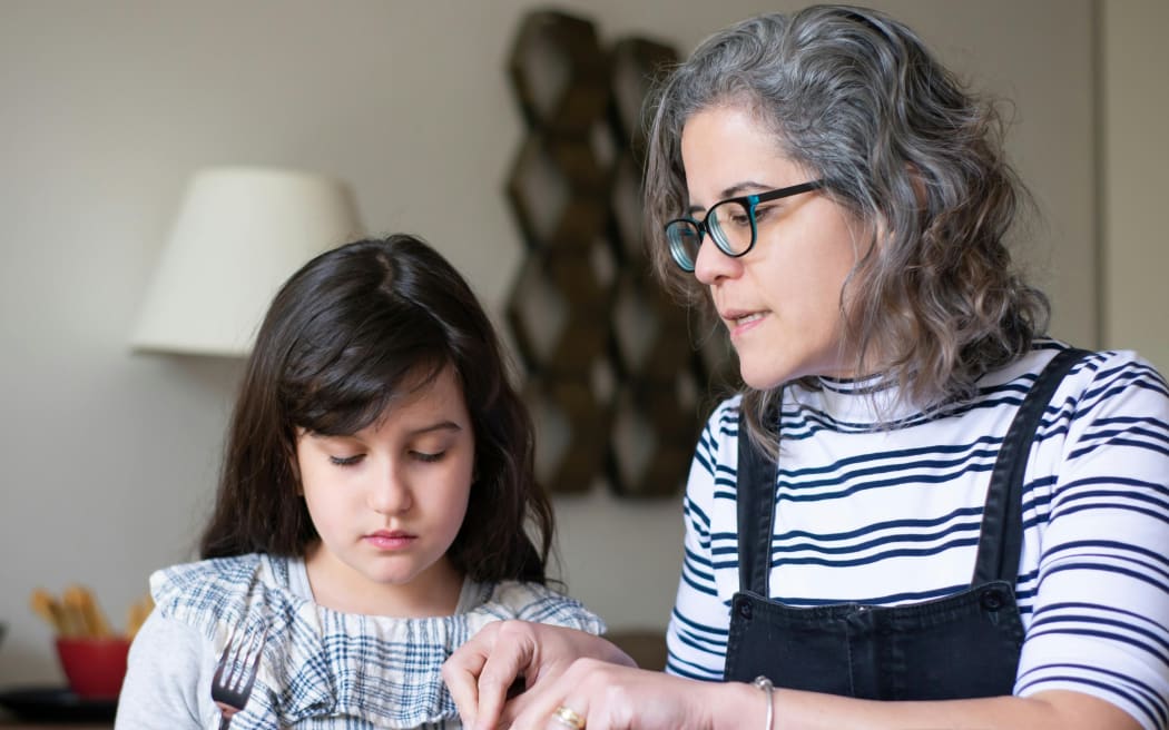 A grey-haired mother sits side by side with a dark haired girl and talks to her.