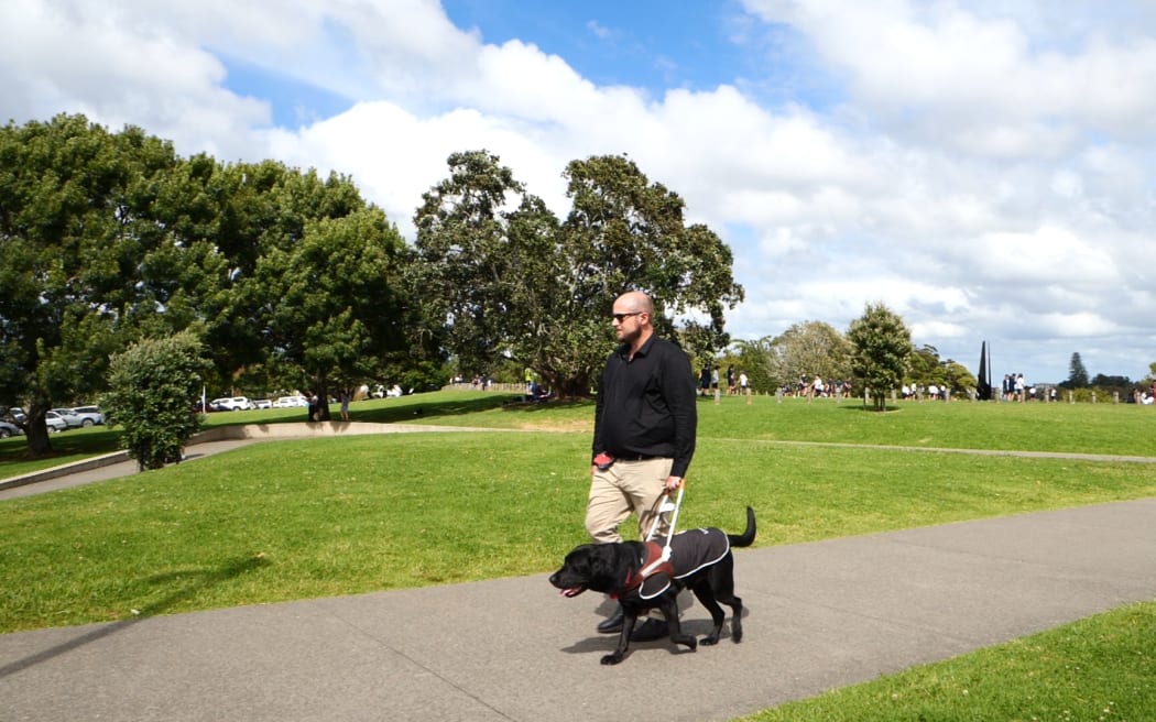 Dan Shepherd and his Guide Dog Ezra take a walk outdoors, in a park with trees in the background.
