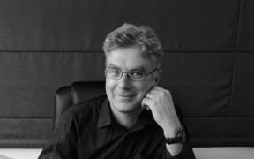 Wellington teacher and author Stephen Tester looks towards the camera with one hand resting on the side of his face. Sitting at a desk with a laptop in front of him.