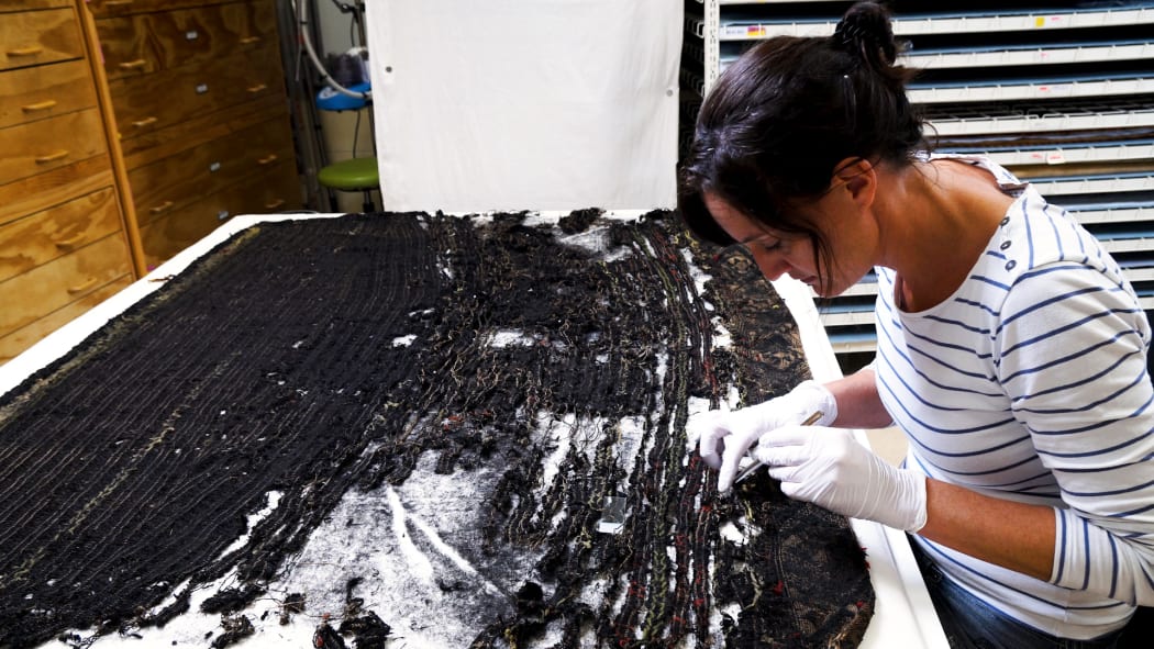 Karin Konold, Te Awe Project Conservator carefully looking at the deterioration of the black muka on a kaitaka (a type of cloak).