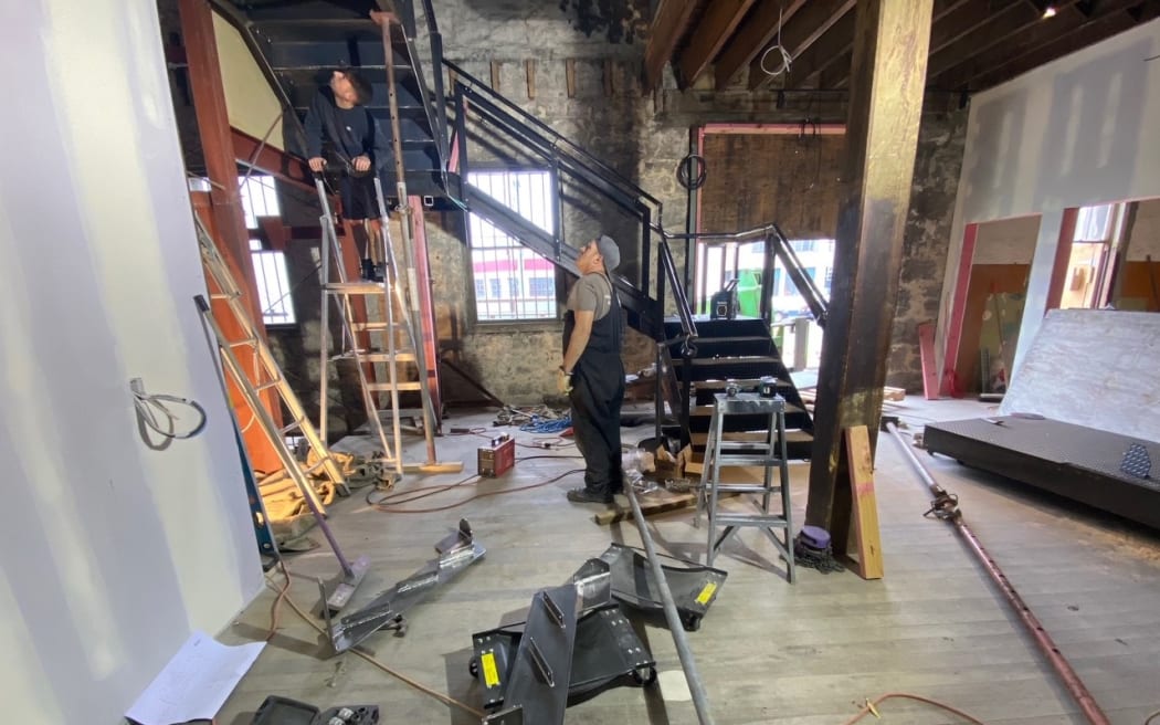 The interior of a heritage building part-way through renovation. Two men confer, one up a ladder and the other looking up at him. There are tools and electrical cables all over the bare floor.