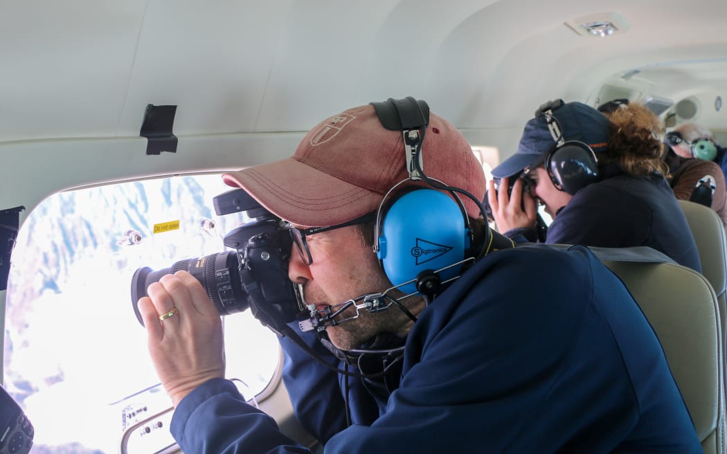 Two people wearing headphones and caps use cameras to take photos looking outside plane windows.