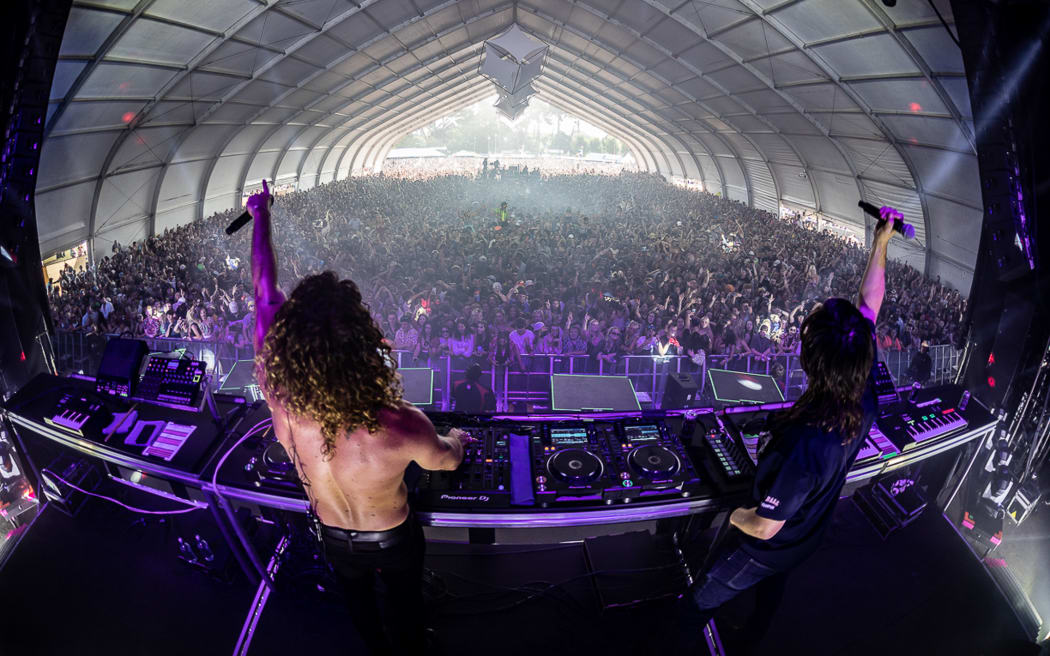 Two DJs holding microphones raise their hands in the air in front of a large crowd.