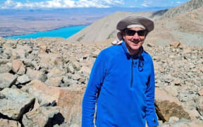 Jamie is wearing a blue half zip fleece and standing on a pile of rocks. It looks like he is on a mountain. Behind him in the distance is a very blue lake further down the mountain.