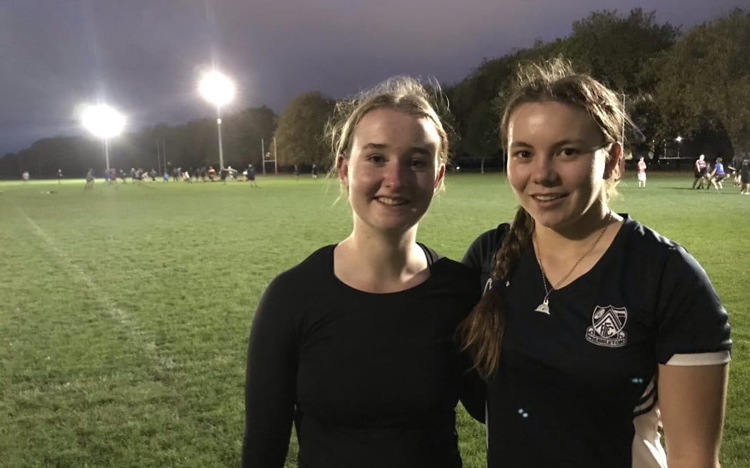 Two girls wearing black stand in front of a sports playing field at dusk with a floodlight shining