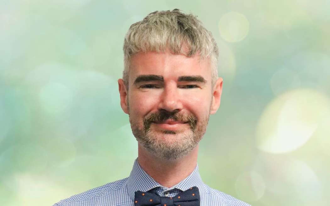 Scott stands in front of a soft green background. He wears a check shirt and a bowtie. He smiles.
