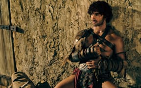 Jordi Webber, wearing a gladiator costume, leans against a wall with his legs open.