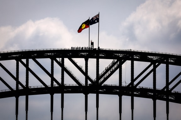 (FILES) Tourists stand under an Aboriginal and Australian national flag located on the top of the Sydney Harbour Bridge on September 27, 2023, ahead of the upcoming "Voice" referendum. A referendum aimed at elevating the rights of Indigenous Australians has instead triggered a torrent of racist slurs and abuse, with toxic debate spreading online and in the media. The October 14 vote will decide whether to finally recognise First Nation peoples in the constitution as Australia's first inhabitants. (Photo by DAVID GRAY / AFP)