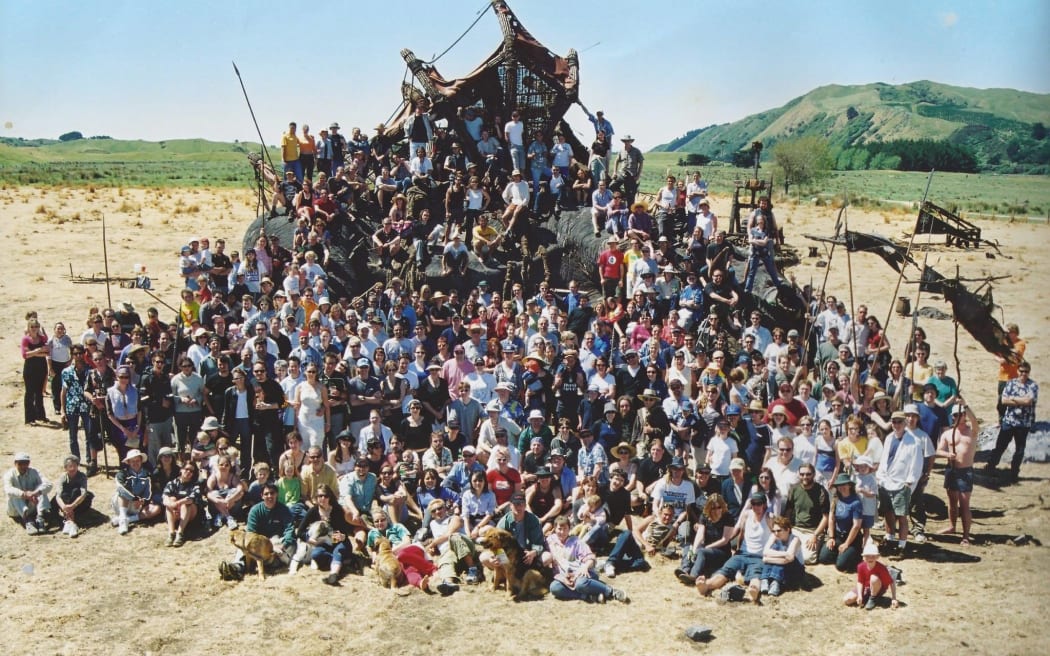 A large group of people, at least 100, standing on an outdoor film set. The ground is sandy, and the group is staggered up a cliff face, with flags sticking out of the fake cliff.