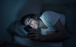 Addicted man chatting and surfing on the Internet with smart phone late at night in bed. Bored, sleepless and tired in dark room with moody light. In insomnia and mobile addiction concept.