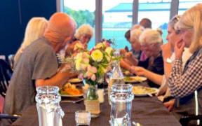 A group of people sitting around a dinner table, with food and drink and flowers. The Dinner Club meets every week in Napier, bringing together people affected by Cyclone Gabrielle for food and company, funded with support from donations and the mayoral relief fund.