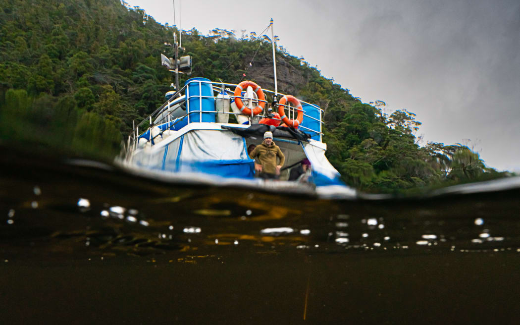 A shot taken half-underwater, half above-water, showing a yellow contraption beneath the surface in dark water. Above the surface, a blue and white boat with orange liferings and a person standing on the back with arms on hips beneath a dark moody sky and a steep slope of native bush.