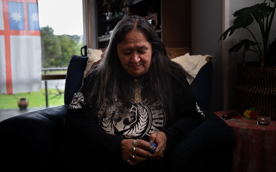 Joyce Harris, photograhed during an interview with RNZ in her living room. She sits in an armchair, holding a blue bandana tightly in her hands.