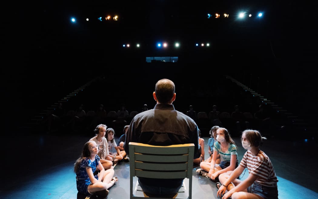 A man sits in a chair on a stage with his back to the camera. Children surround him in a U-shape on the floor.