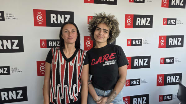 Two women stand against a wall that reads RNZ.