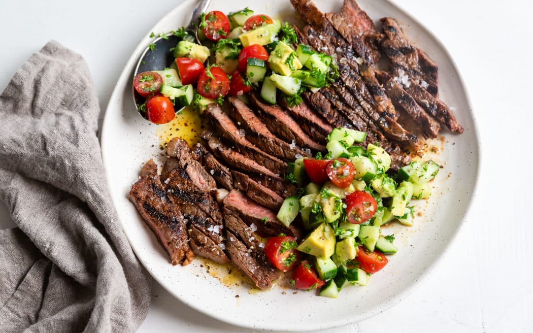 A round white plate showing sliced steak and cucumber and avocado salad, with a grey linen serviette.