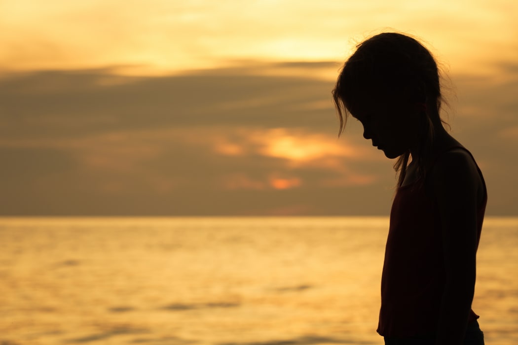 Portrait of sad blond little girl standing on the beach at sunset time.