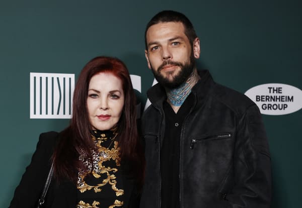 Priscilla Presley, with long auburn hair, stands next to Navarone Garibaldi who has a beard and a tattooed neck.