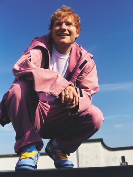 British pop star Ed Sheeran smiling, kneeling down in a pink outfit.
