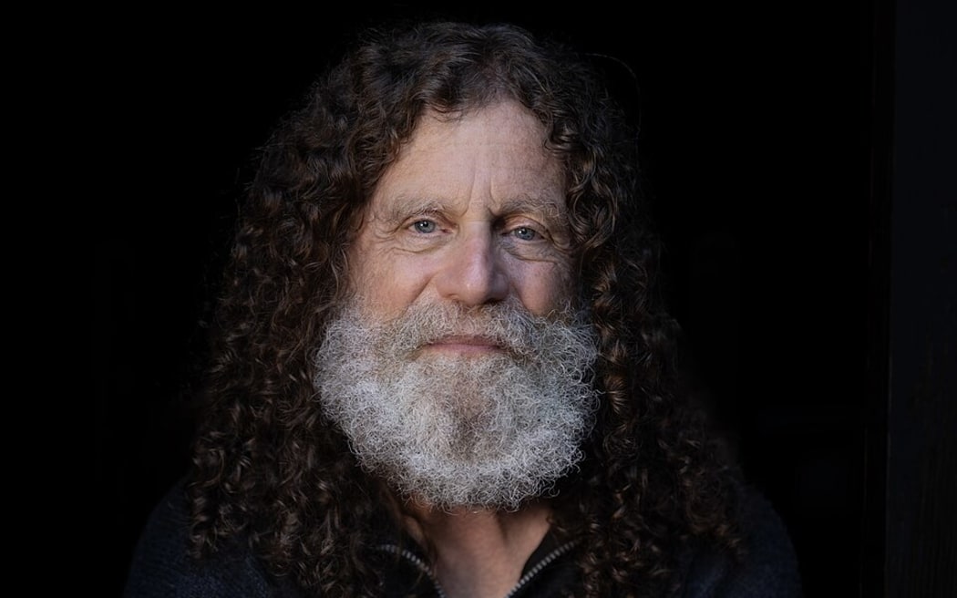 Robert Sapolsky has long brown hair and a white beard and sits on a stool holding what appears to be a human skull.