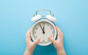 Baby hands holding white alarm clock on light blue table background. Pastel color. Time concept. Closeup. Point of view shot. Top down view.