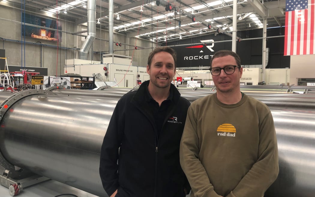 Ben and Dylan smile at the camera. They are standing in front of rows of large metallic tubes in a huge warehouse with the ROCKET LAB logo on the back wall. An American flag is hung behind them.