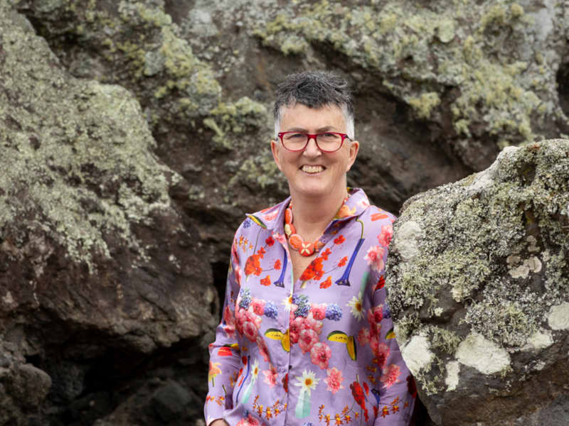 Dr Lucy O'Hagan - a woman with short grey hair and red glasses - stands smiling between some large rocks.
