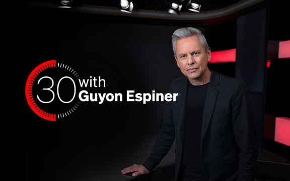 Guyon Espiner on a black background, looking at camera with logo behind him