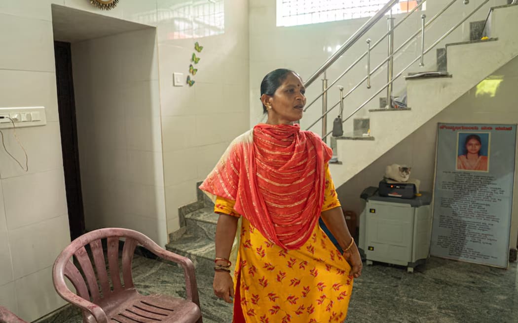 An Indian woman in a bright orange and red saree standing inside next to a staircase, red chair and image of her daughter.