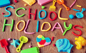 the text school holidays made from modelling clay of different colors and some beach toys such as toy shovels and sand moulds, on a rustic wooden surface