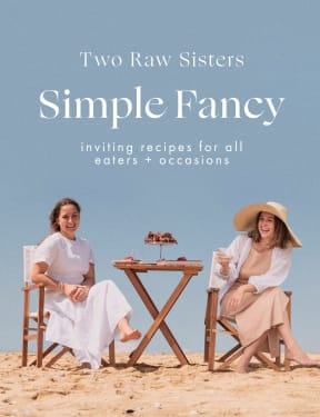 Cover of Two Raw Sisters' cookbook called "Simple Fancy" which shows a picture of The Two Raw Sisters wearing summer dresses sitting on folding chairs beside a folding table on a beach.